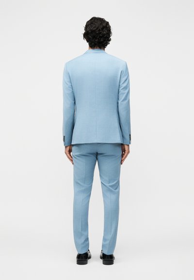 Άνδρας με καστανά σγουρά μαλλιά, ντυμένος με ανοιχτού μπλε κοστούμι και μαύρα παπούτσια, στέκεται με την πλάτη στραμμένη σε λευκό απλό φόντο.