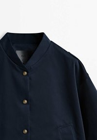 Veste bleu marine avec col mandarin, boutons dorés et une poche latérale unique. Fabriquée en tissu lisse et texturé avec une finition légèrement brillante.