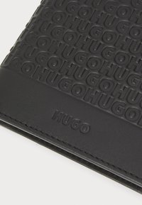 Portofel negru cu marca „HUGO” embosată, având o textură fină și un design elegant, evidențiind detalii cu litere îngroșate.