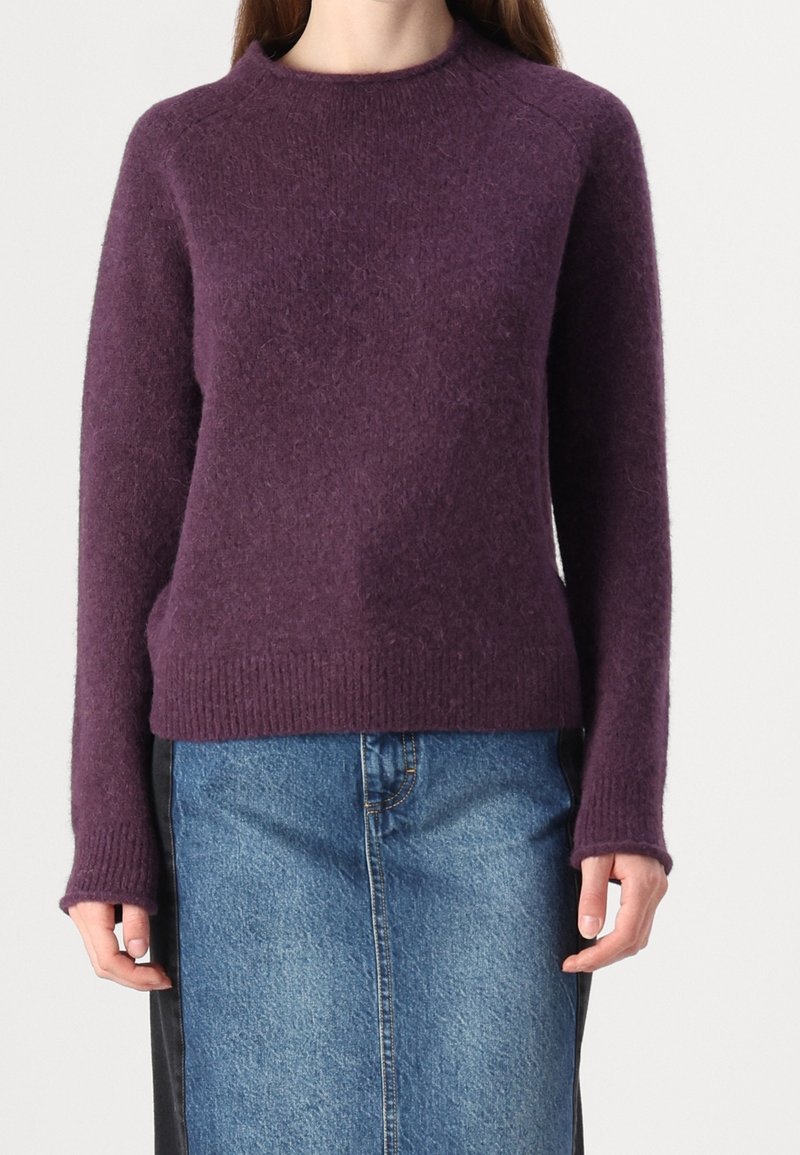 Femme portant un pull douillet à manches longues violet, associé à une jupe en denim bleue avec des panneaux latéraux noirs, debout devant un fond uni.