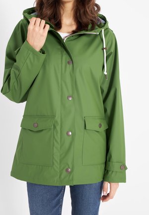 Femme portant un imperméable vert à capuche avec des poches boutonnées et un cordon blanc, associé à un jean bleu, sur fond blanc.