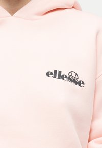 Ανοιχτό ροζ φούτερ με κράνος και μαύρο λογότυπο "ellesse" και ένα ημικύκλιο γραφικό στο στήθος, που δείχνει το ανώτερο μέρος του κορμού και την περιοχή της κουκούλας.