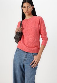 Pull en tricot corail vif avec ourlet et poignets côtelés, associé à un jean en denim bleu ample. Le mannequin tient un petit sac noir.