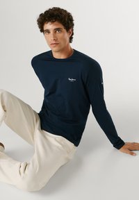 Ung mand med krøllet hår iført en marineblå langærmet skjorte og beige bukser, siddende på gulvet foran en ensfarvet baggrund.