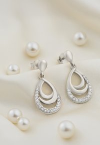 Orecchini a goccia d'argento con design a strati, caratterizzati da pietre trasparenti lungo il bordo inferiore, posizionati su un tessuto morbido e chiaro con perle.