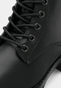 Gros plan sur une botte en cuir noir avec des œillets métalliques ronds et des lacets noirs serrés, montrant une texture lisse et des détails de couture.
