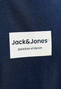 Jack & Jones brand logo and tagline "Definition of Denim" printed in black on a white rectangular label on navy blue fabric.