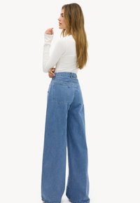 Mujer con camiseta blanca de manga larga y vaqueros azules de tiro alto y piernas anchas, de pie de perfil con los brazos cruzados.