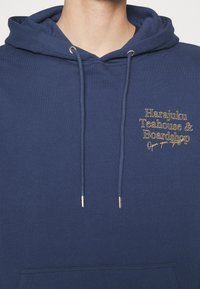 Felpa blu navy con cappuccio, dotata di cordini, e testo giallo sul petto che recita "Harajuku Teahouse & Boardshop Aperto su richiesta."