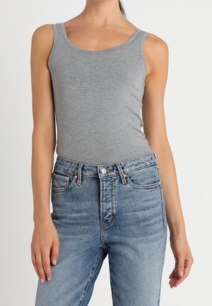Femme portant un débardeur gris ajusté et un jean bleu taille haute, debout devant un fond clair uni, seule la partie supérieure du corps est visible.