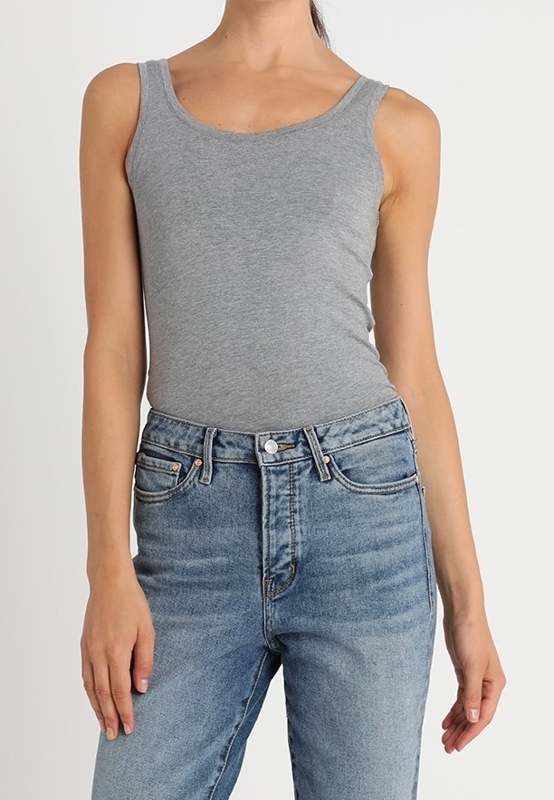 Femme portant un débardeur gris ajusté et un jean bleu taille haute, debout devant un fond clair uni, seule la partie supérieure du corps est visible.