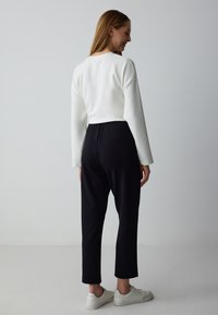 Weißer, verkürzter Sweatshirt mit langen Ärmeln, kombiniert mit schwarzen, elastischen Hosen. Das Model trägt hellfarbene Sneaker. Minimalistisches Design, glatte Textur.