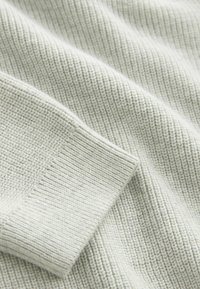 Tessuto grigio chiaro con texture che presenta un motivo a coste, con un dettaglio a polsino visibile e una finitura morbida e tessuta.