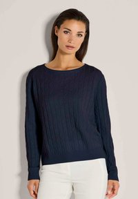 Frau, die einen navyblauen Kabelstrickpullover und weiße Hose trägt, steht vor einem schlichten, hellbeigen Hintergrund.