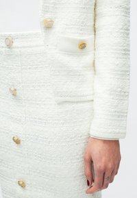 Λευκό σακάκι από τουίντ με υφή, διακοσμημένο με χρυσά κουμπιά και μία τσέπη. Η συνοδευτική φούστα έχει παρόμοια λεπτομέρεια κουμπιών και υφή.