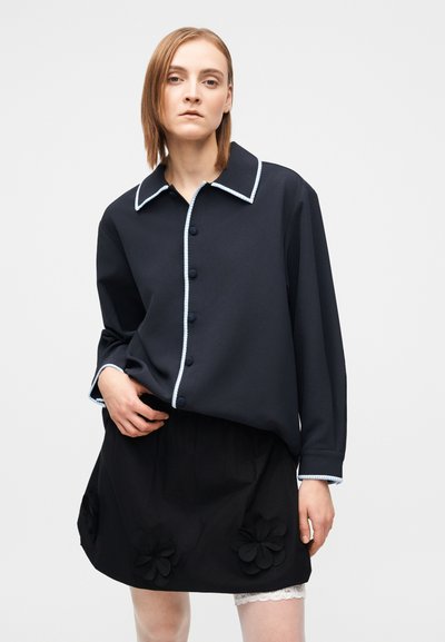 Melnas apkaklītes krekls ar zilu apmali un pogām, kopā ar melnu svārku ar trim ziedu aplikācijām un mežģīņu apmali. Gluda auduma tekstūra.