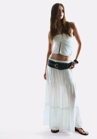 Top halter alb, scurt, cu detalii pliate, asortat cu o fustă maxi albă, fluxată, cu straturi, și un cureaua lată neagră cu ținte argintii.