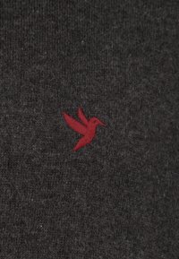 Donkergrijze stof met een subtiele textuur en een klein rood geborduurd kolibrilogo aan de linkerkant.