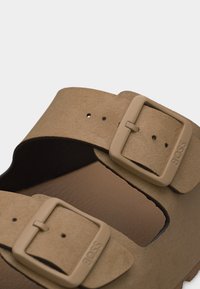 Gros plan sur une sandale beige avec deux sangles ajustables ornées de boucles carrées embossées du logo "BOSS".