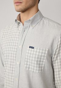 Camisa de cuadros de botón en gris claro y blanco con un bolsillo en el pecho; presenta una etiqueta de logo y una tela texturada.