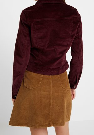 Femme portant une veste en velours côtelé bordeaux foncé et une jupe en velours côtelé marron clair, debout dos à la caméra.