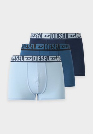 Mężczyzna bez koszulki w granatowych bokserkach Diesel, stoi boso na jednolitym białym tle, patrząc na bok.
