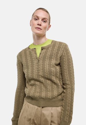 Femme aux cheveux blonds tirés en arrière, portant un pull beige en maille torsadée sur un top vert citron et un pantalon plissé beige sur fond blanc.
