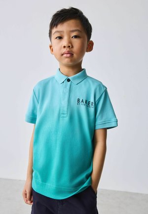 Junger Junge steht mit einer Hand in der Tasche, trägt ein türkisfarbenes Poloshirt mit dem Schriftzug „BAKER BY TED BAKER“ auf der Brust, neutraler Hintergrund.