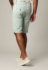 Man in lichtgroene opgerolde shorts, gebloemd shirt gestopt in de broek, en witte instappers, staande tegen een effen achtergrond.