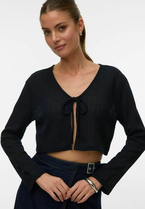 Kvinde med lys hud og brunt hår samlet bagpå, iført en sort kort cardigan med bindebånd foran og mørke bukser med højt talje og et sølvarmbånd.