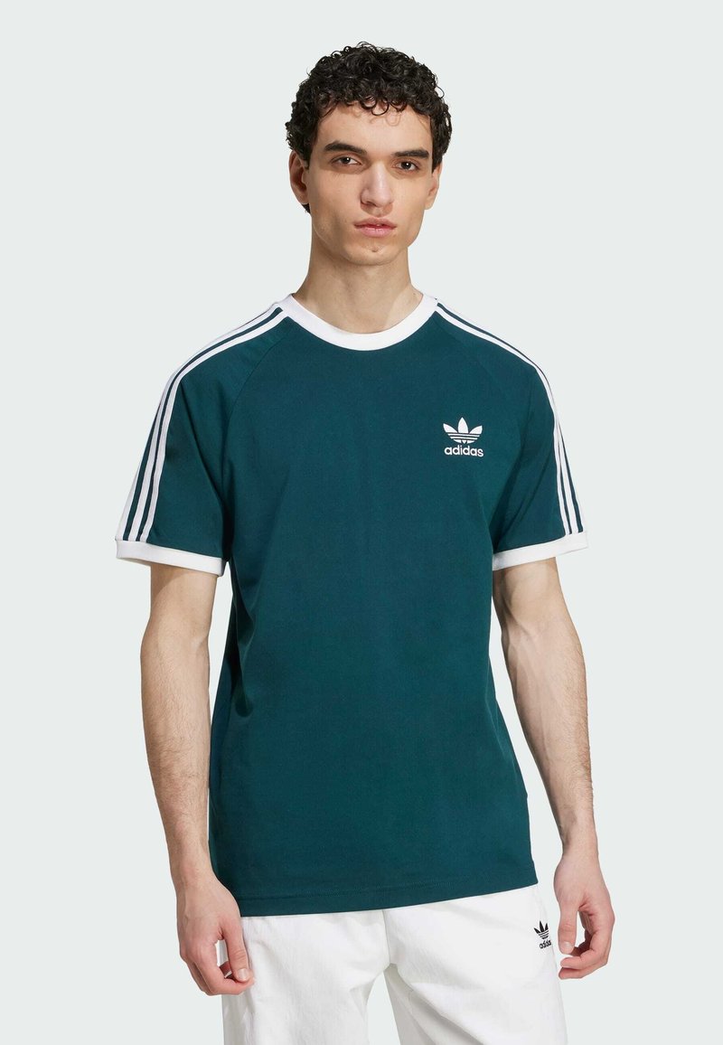 T-shirt en coton vert foncé avec un col ras du cou et des ourlets de manches blancs, présentant trois rayures verticales blanches sur les épaules et un petit logo sur la poitrine.