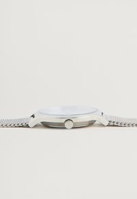 Ατσάλινο ρολόι με επίπεδη στρογγυλή κάσα, λευκό καντράν και υφασμένη μπάντα. Περιλαμβάνει ένα μικρό κορώνα για ρύθμιση της ώρας.