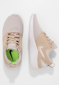 Beige träningsskor i mesh och syntetmaterial, med en kontrasterande grön innersula och vit branding. Rundad tå och vadderad sula.