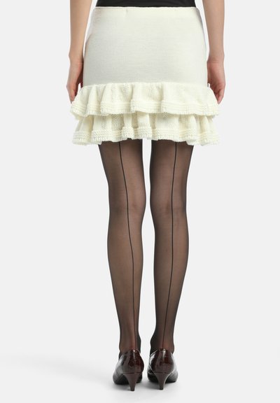 Valkoinen neulemini-hame, jossa on röyhelösisustus, yhdistettynä läpikuultaviin mustiin sukkahousuihin, joissa on pystysauma, ja ruskeisiin kärkitossuihin.