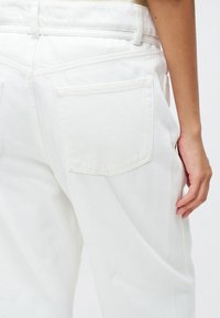 Hvite denimbukser har høy midje, sidelommer og to baklommer, med synlige sømmer på sømmene.