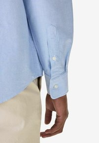 Vaaleansininen kauluspaita, jossa on pisteriipinystykset ja näkyvät saumat hihoissa, yhdistettynä beigen värisiin housuihin. Hieman havaittavaa tekstuuria.