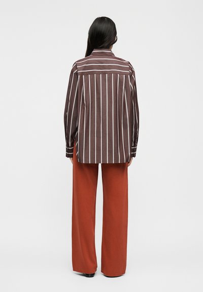 Γυναίκα με μακριά σκούρα μαλλιά, φορώντας καφέ και λευκό πουκάμισο με κάθετες ρίγες και φαρδιά παντελόνια σε σκουριά απόχρωση, στέκεται με την πλάτη προς το μέρος μας.