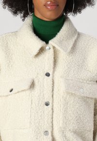 Cream-colored textured jacket with a collared neck, two front pockets, and silver snap buttons. Worn over a green turtleneck.