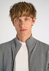 Grå zip-up jacka med ett texturerat mönster, hög krage och stickad tyg. Bärs över en enkel vit T-shirt.