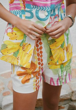 Ruce zapínající barevnou bundu s květinovým a geometrickým vzorem a odpovídající kraťasy, na pravém zápěstí stříbrné náramky.