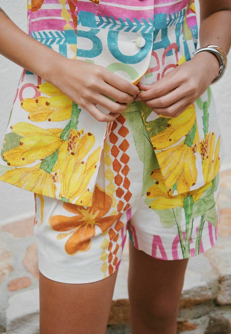 Des mains boutonnant une veste et un short assortis à motifs floraux et géométriques colorés, avec des bracelets argentés au poignet droit.