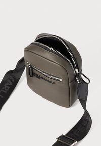 Borsa in pelle verde oliva a forma quadrata con cerniere argentate, tasca frontale e tracolla nera regolabile con logo. Superficie texturizzata con dettagli.