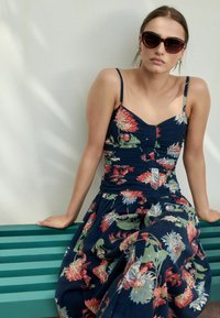Blumendruckkleid aus navy Stoff mit Spaghettiträgern. Mit gerafften Details am Oberteil und einem fließenden Rock. Dunkle Sonnenbrille.