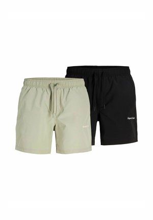 Junger Mann mit kurzen blonden Haaren steht barfuß in hellbeigen Badeshorts vor einem schlichten weißen Hintergrund.