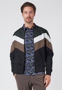 Giacca bomber color block con pannelli verdi, bianchi, marroni e neri. Tessuto liscio, polsini a costine e chiusura con zip. Camicia floreale sotto.