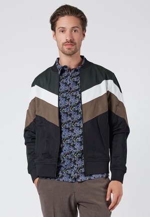 Color-blocked bomber jacket featuring green, white, brown, and black panels. Smooth fabric, ribbed cuffs, and a zip closure. Floral shirt underneath.