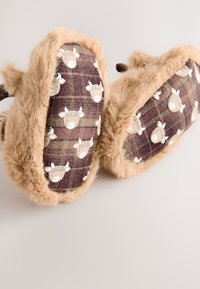 Zapatillas peludas marrones con un patrón de cuadros que presenta diseños de vacas escocesas blancas. Material suave y texturizado con un borde mullido y forma redondeada.