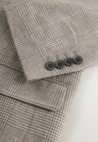Rukáv béžovo-hnedého kockovaného saka so štyrmi tmavými gombíkmi a malým vreckom na neutrálnom pozadí.
