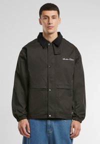 Black jacket with a textured collar, snap buttons, two front pockets, and embroidered logo on the left chest. Elastic cuffs.