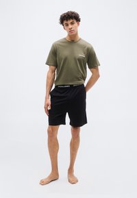 Νέος άνδρας με σγουρά μαλλιά, όρθιος και ξυπόλητος, φοράει ελαφρώς χακί κοντομάνικη μπλούζα και μαύρο σορτς με branded ζώνη στη μέση.